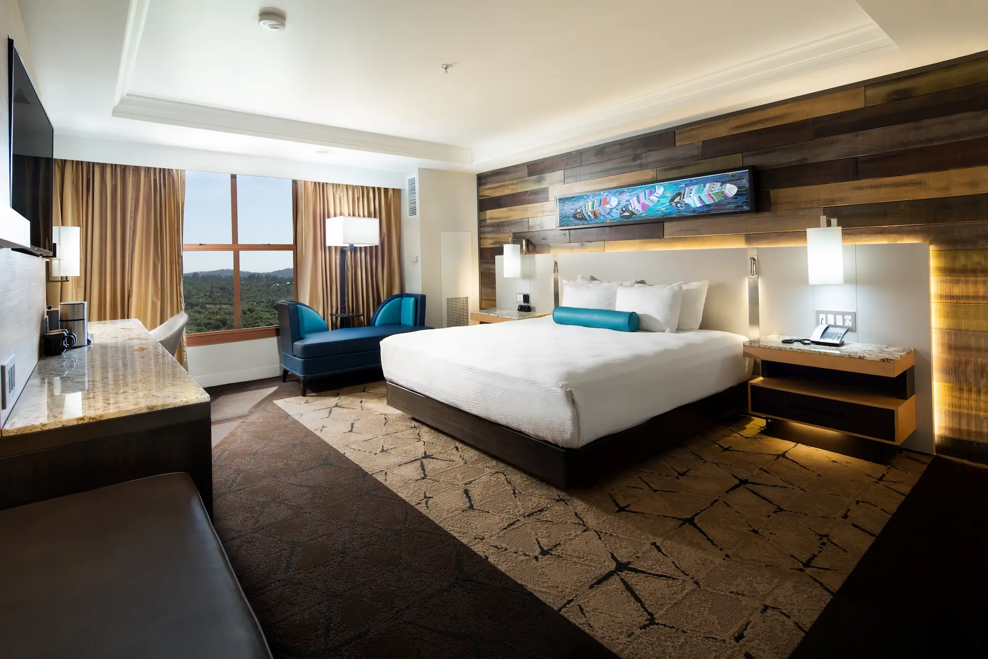 A modern hotel room with a large king-sized bed in the center, adorned with white linens and a turquoise bolster pillow. The headboard features a wooden panel design with ambient lighting and a colorful abstract painting above it. On either side of the bed are nightstands; the right one holds a phone. A large window with beige curtains shows a scenic green landscape outside. Near the window is a dark blue bench with two matching chairs and a floor lamp. To the left is a marble-topped desk with a flat-screen TV mounted above it. The carpet is a mix of brown and beige with a geometric pattern.