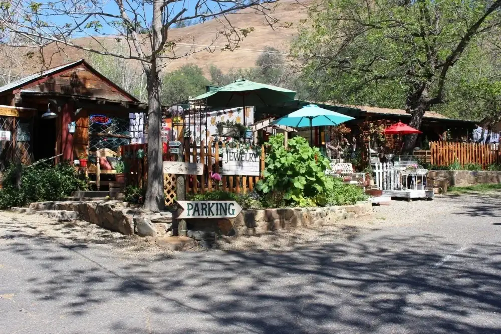 A rustic outdoor scene featuring a small, wooden building on the left with a sign that reads "OPEN" in bright neon colors. In front of the building, a wooden fence displays another sign that says "JEWELRY." To the right, there are various items displayed under colorful umbrellas, including teal and red ones, suggesting a market or crafts area. A larger wooden sign in front points to "PARKING" with an arrow. The area is surrounded by greenery, trees, and potted plants, with a background of rolling hills under a blue sky.